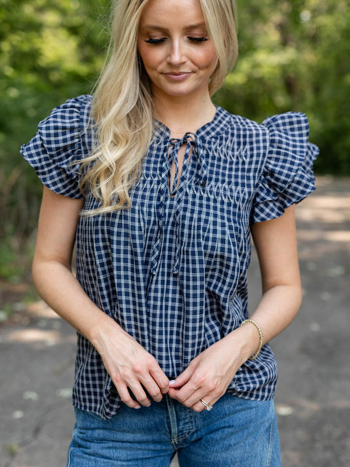 A woman wearing a navy and white gingham check short-sleeve blouse with ruffled sleeves and a tie neckline, paired with blue jeans, standing outdoors in front of a blurred green background.