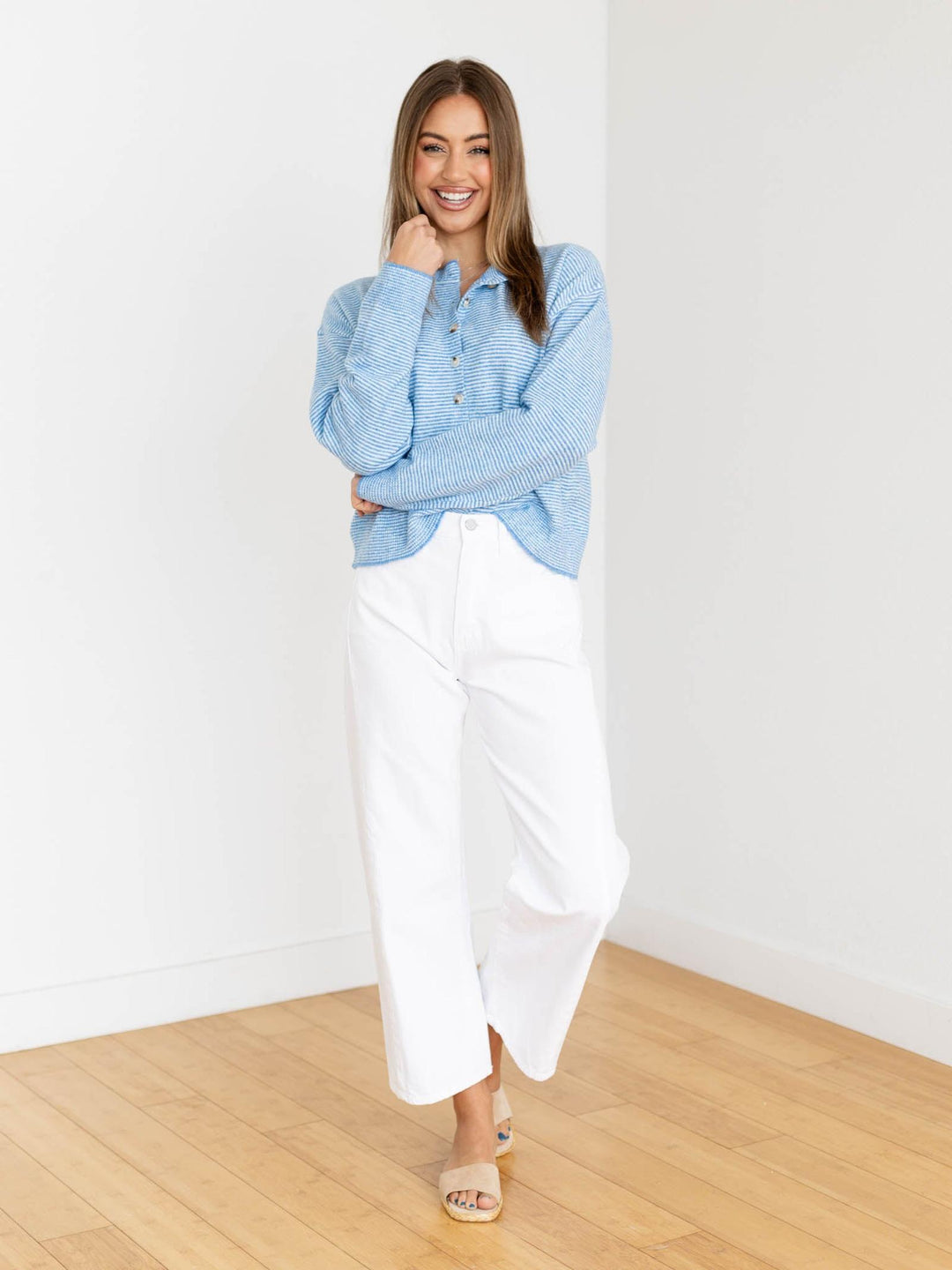 Woman wearing a blue sweater and white pants standing in a room with wooden floor and white walls.