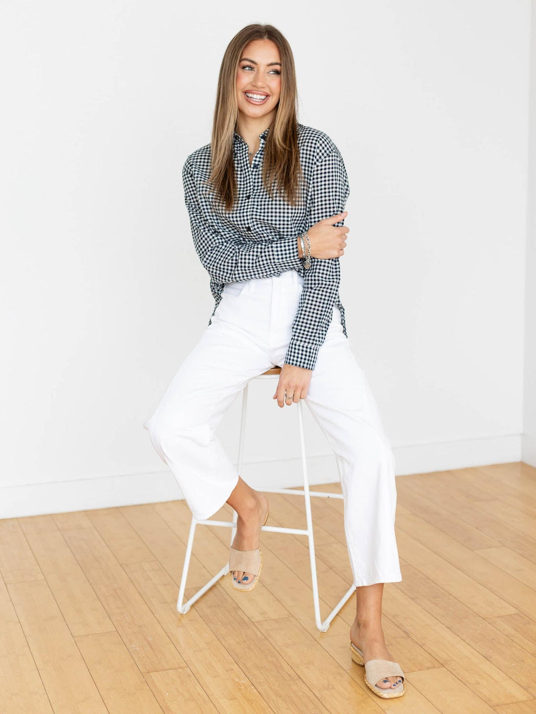 Woman wearing a checkered shirt and white pants sitting on a stool against a white wall.