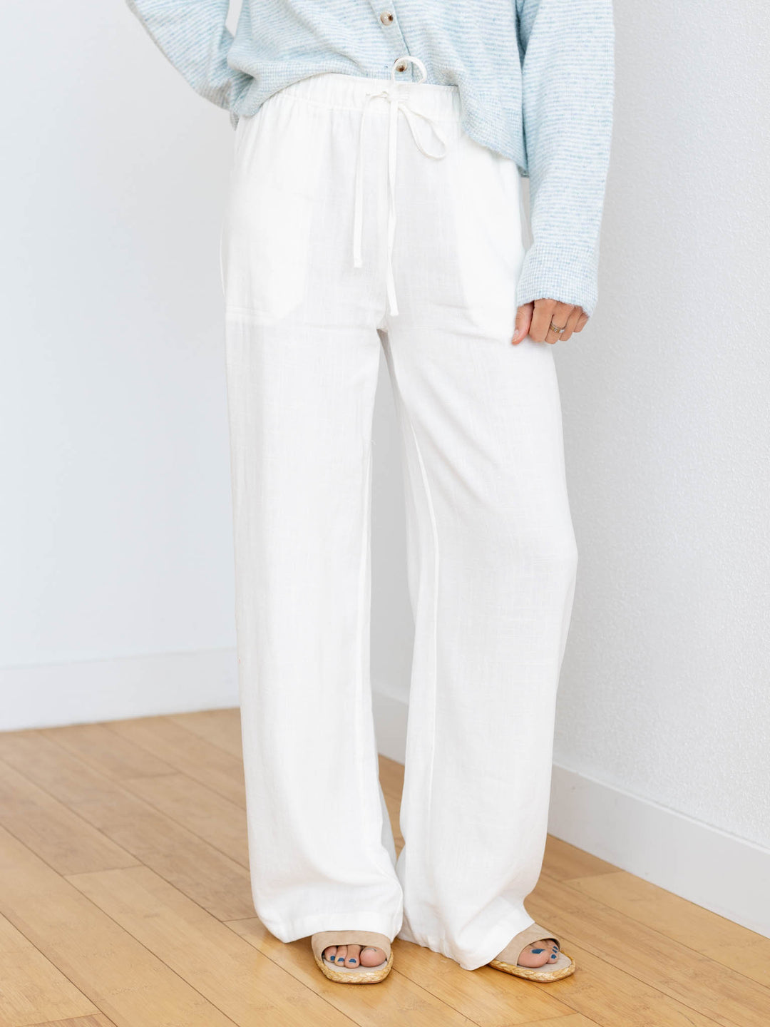 Person wearing light blue shirt and white wide-leg pants standing on a wooden floor.