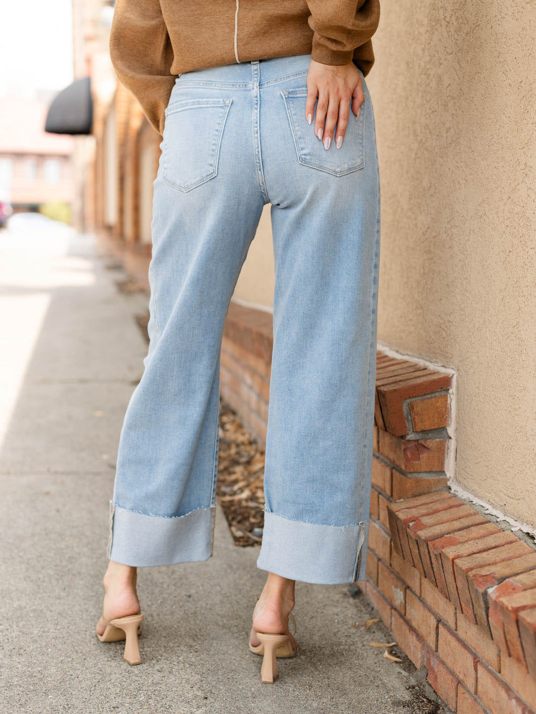 A person shown from the back wearing light-wash wide-leg cropped jeans with rolled cuffs, a brown cropped sweater, and beige heeled sandals, standing outdoors beside a brick wall and sidewalk.
