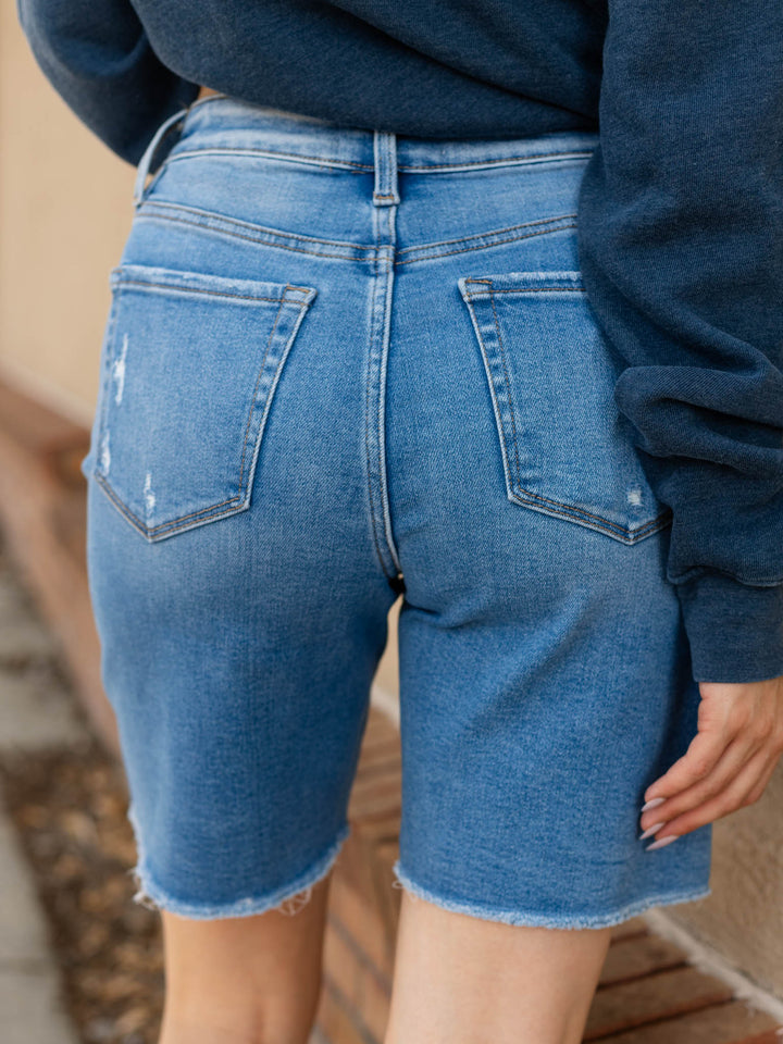Rear view of a person wearing light-to-medium wash denim shorts with a frayed hem and back pocket details, paired with a dark blue sweatshirt.