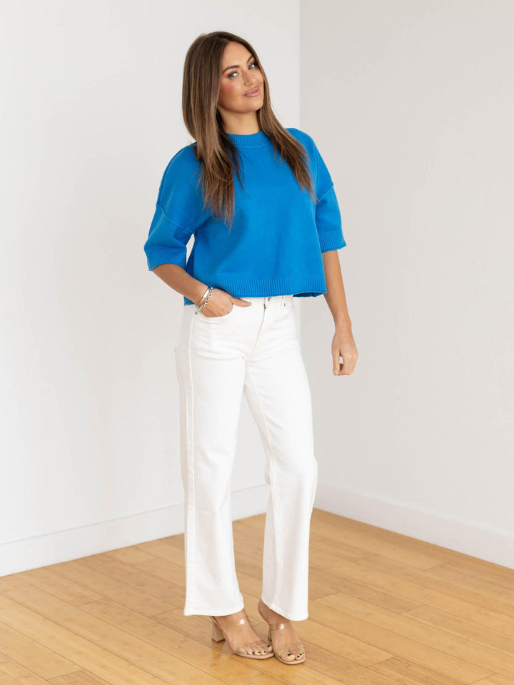 Woman wearing a blue top and white pants standing in a room with wooden floor and white walls.