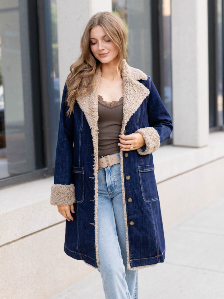 A woman wearing a navy blue denim-style coat with beige sherpa lining, styled over a brown top and light blue jeans, standing outdoors in front of a modern building.