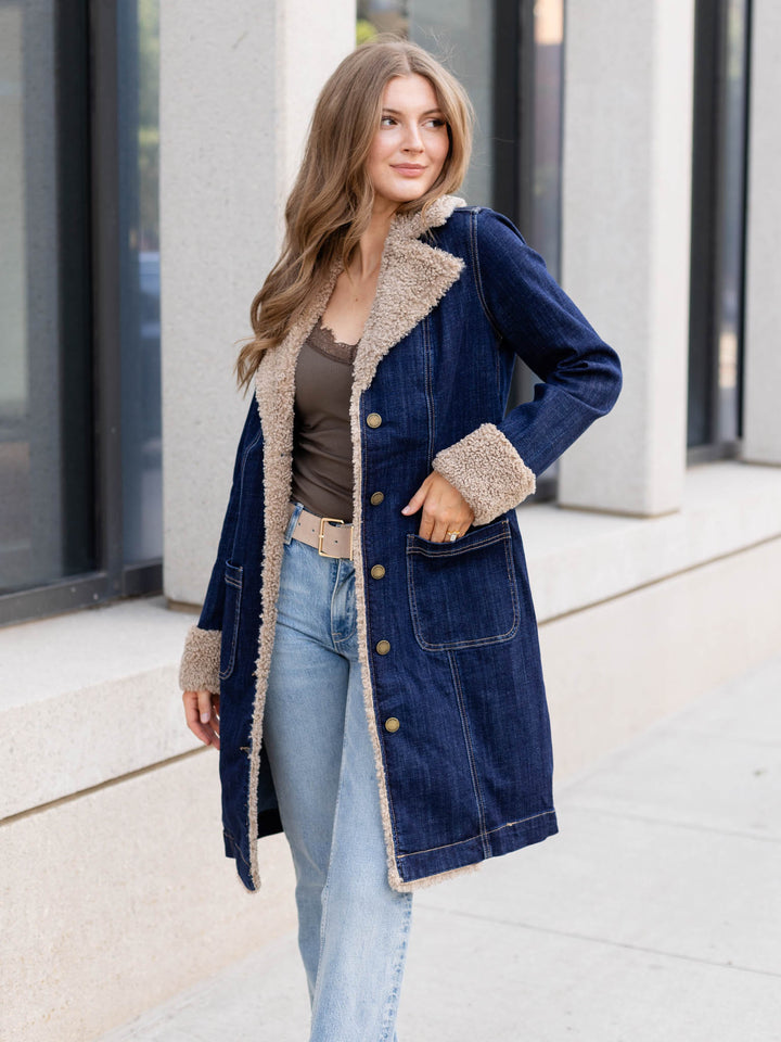 A woman wearing a dark blue denim coat with beige sherpa lining, styled with a brown top and light blue jeans, standing outdoors in front of a modern building.