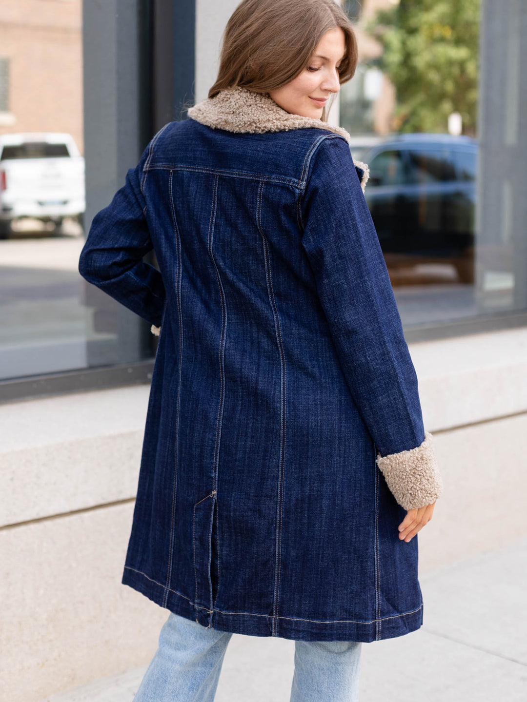 A woman wearing a dark blue denim coat with beige sherpa lining on the collar and cuffs, photographed outdoors on a sidewalk.