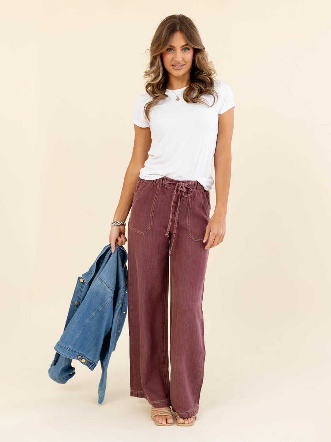 Woman wearing a white t-shirt and maroon pants holding a denim jacket on a beige background