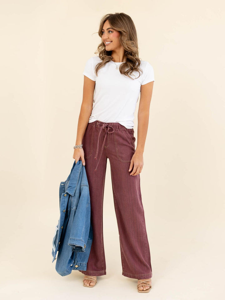 Woman wearing a white t-shirt and maroon wide-leg pants, holding a blue denim jacket on a beige background.