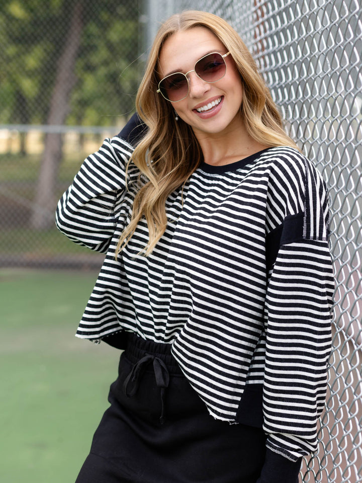 A woman wearing a black and white striped long-sleeve crop top and black pants, leaning against a chain-link fence outdoors.