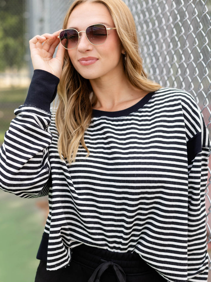 A woman wearing a black and white horizontally striped long-sleeve top with solid black cuffs, standing in front of a chain-link fence.