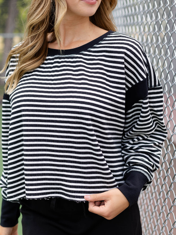 A woman wearing a black and white horizontally striped long-sleeve top with solid black ribbed trim at the crew neck, cuffs, and shoulders, standing in front of a chain-link fence.