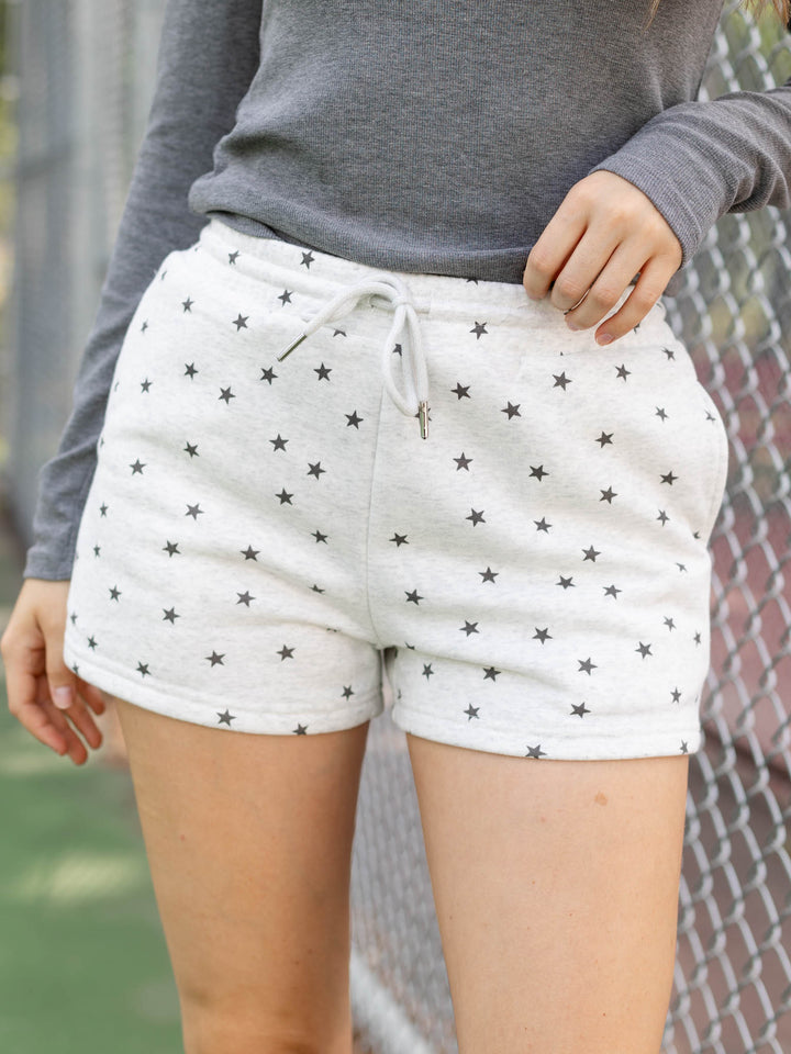 A person wearing light heather gray drawstring shorts with a black star pattern, paired with a gray long-sleeve top, standing in front of a chain-link fence.