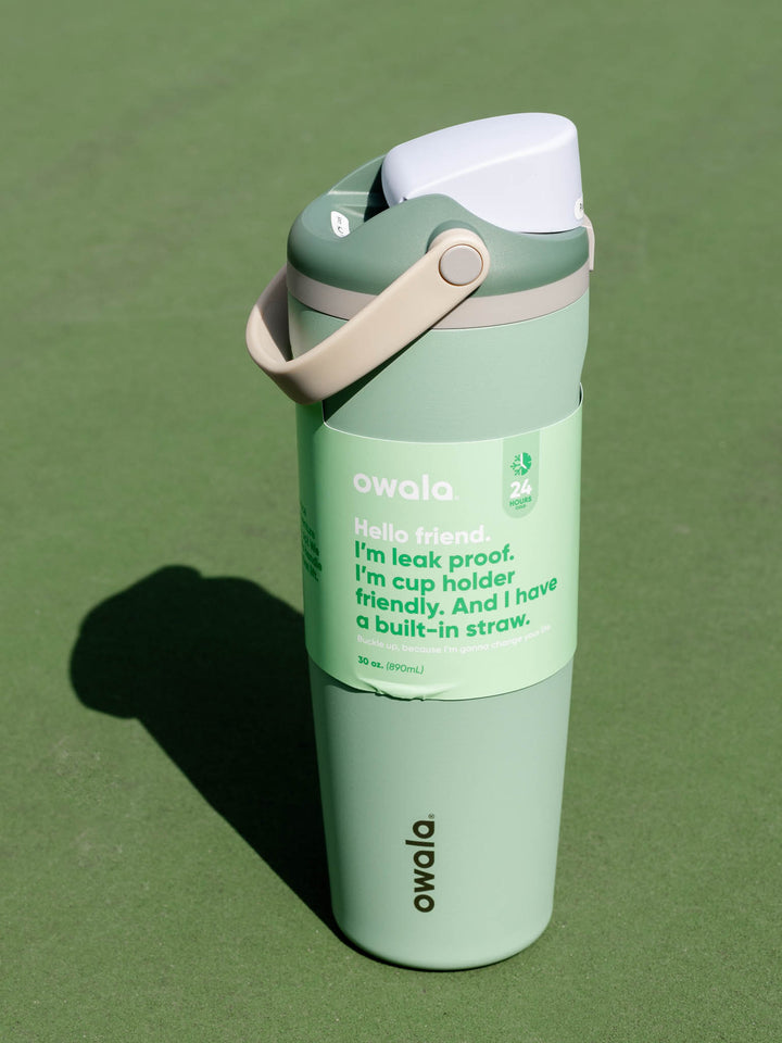 A Owala leak-proof travel tumbler in sage green with a white lid and beige carrying strap, standing on a green surface.