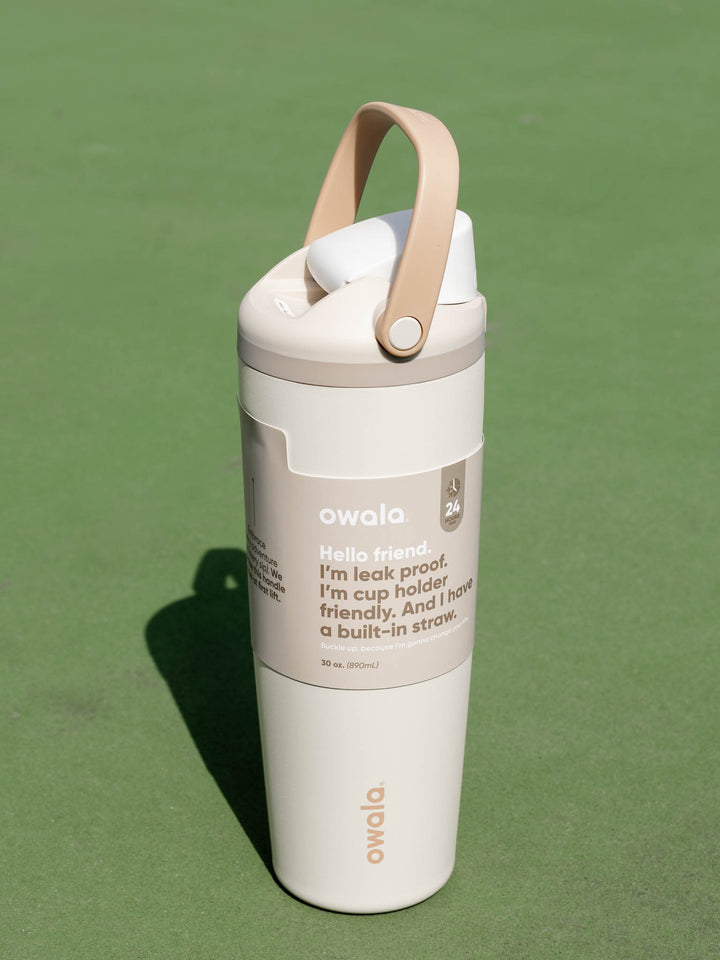 An Owala insulated travel tumbler with a beige carry loop and a white/light beige body, standing on a green surface.