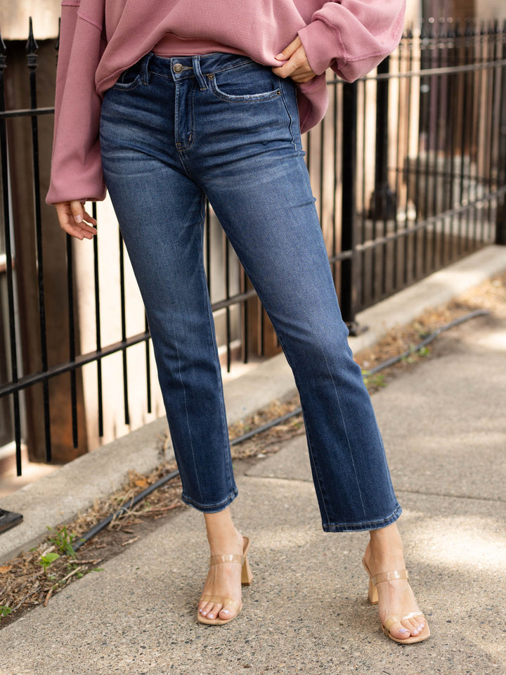 A person wearing dark blue cropped straight-leg jeans, a dusty pink sweatshirt, and clear high-heeled sandals standing on a sidewalk next to a black metal fence.
