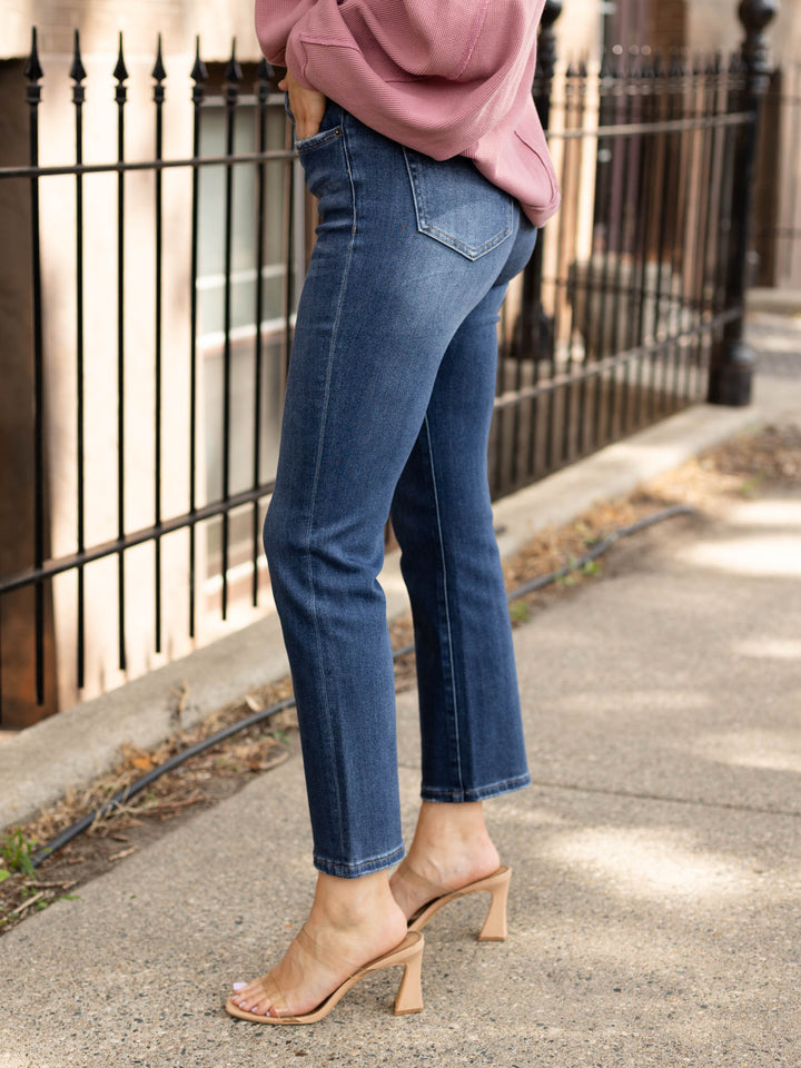 A person wearing blue straight-leg jeans, a dusty pink sweatshirt, and beige heeled mule sandals, standing on a sidewalk next to a black metal fence.
