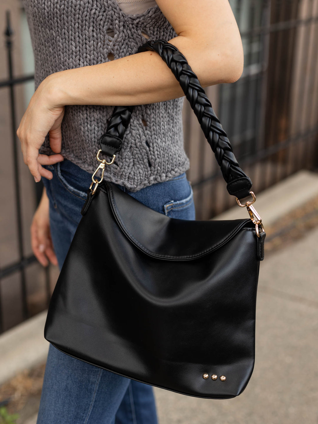 A person wearing a gray knit sweater and blue jeans carries a large black leather shoulder bag with a braided black strap and gold-tone hardware.