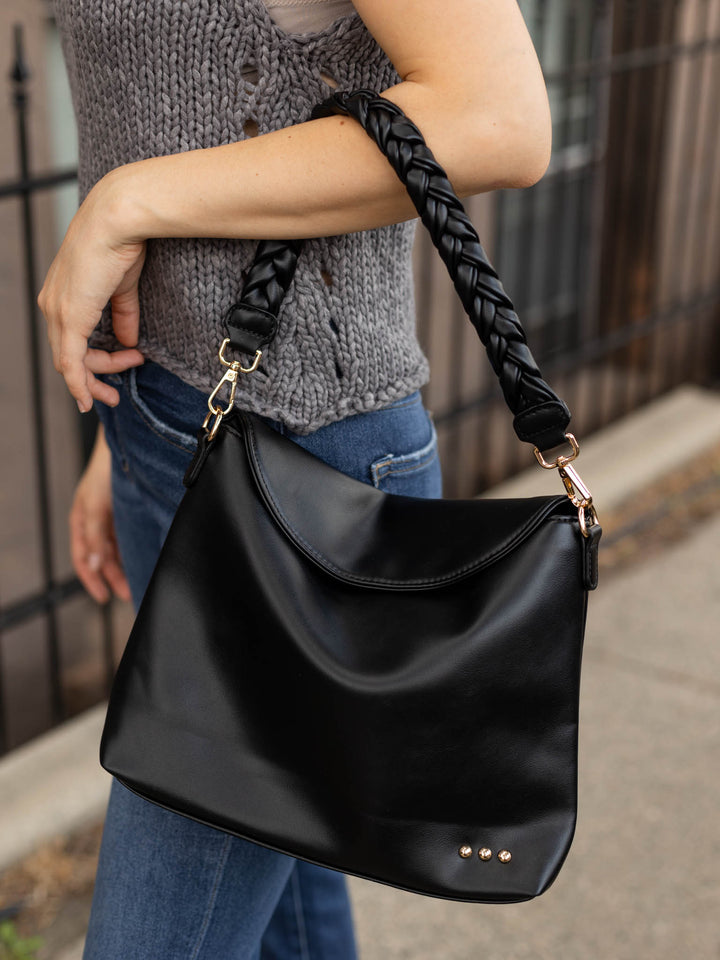A person wearing a gray knit sweater and blue jeans carries a large black leather shoulder bag with a braided black strap and gold-tone hardware.