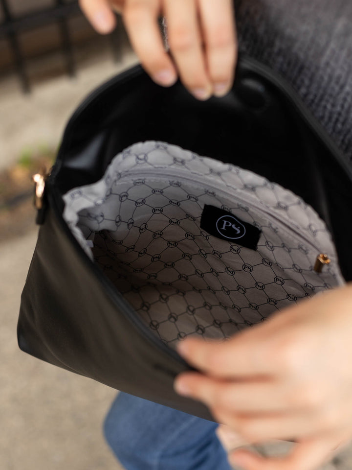 Close-up of a person holding open a black shoulder bag to show the interior lining, which features a gray and black geometric pattern and a small black brand patch.