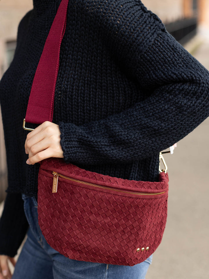 A person wearing a dark navy knit sweater and blue jeans carries a burgundy woven crossbody bag with a matching burgundy strap and gold-tone hardware.