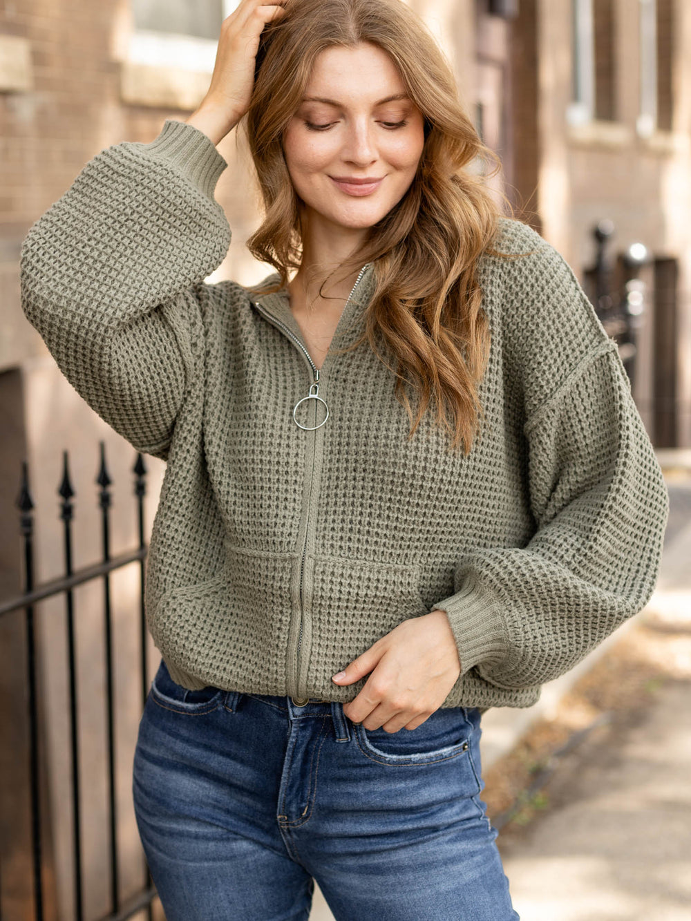 A woman wearing an oversized waffle-knit zip-up sweater in sage green with blue jeans, standing outdoors on a street with a blurred background.