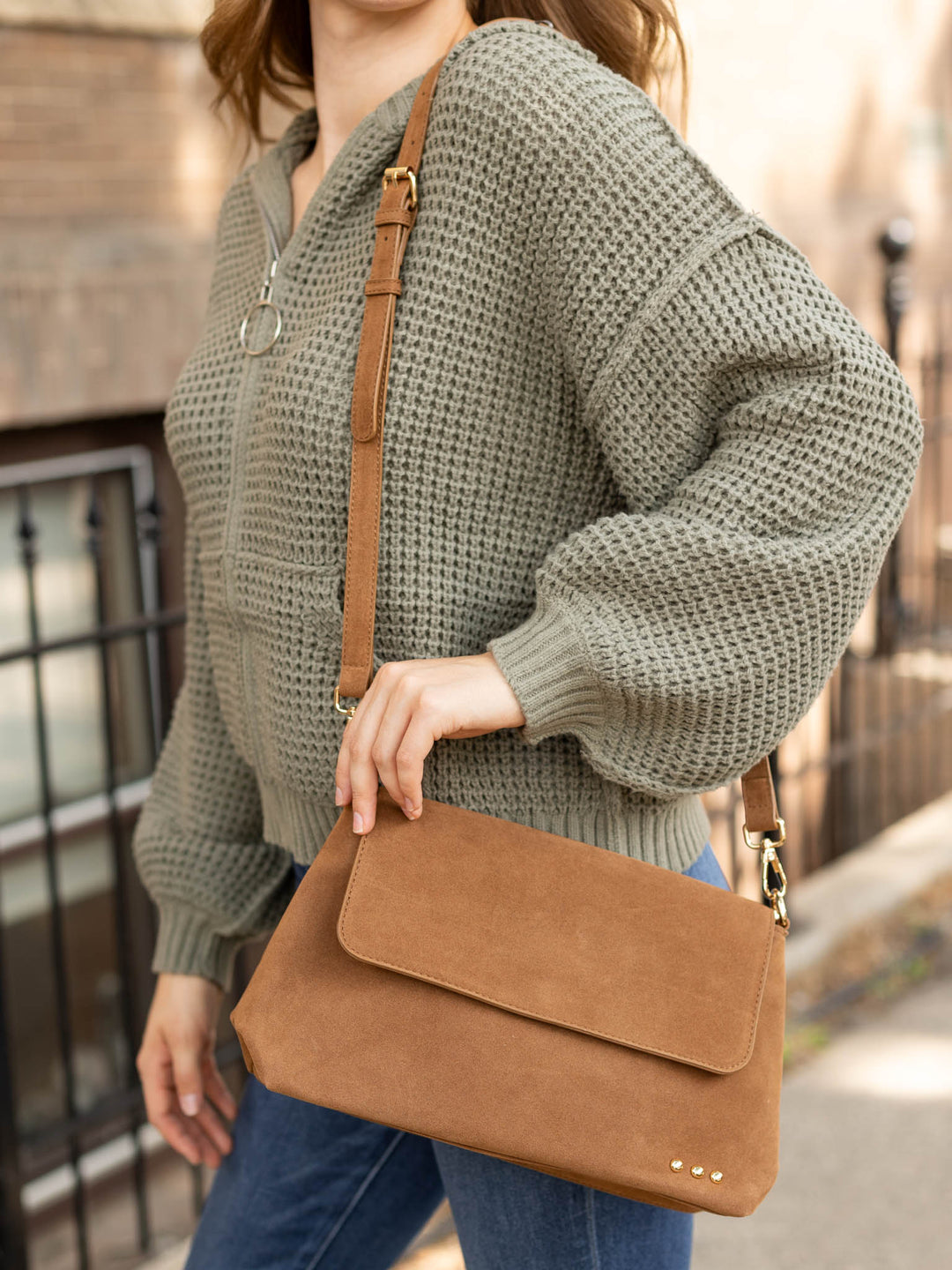 A woman wearing a chunky knit olive green sweater and blue jeans carries a tan suede crossbody bag with a brown strap and gold-tone hardware in an outdoor urban setting.