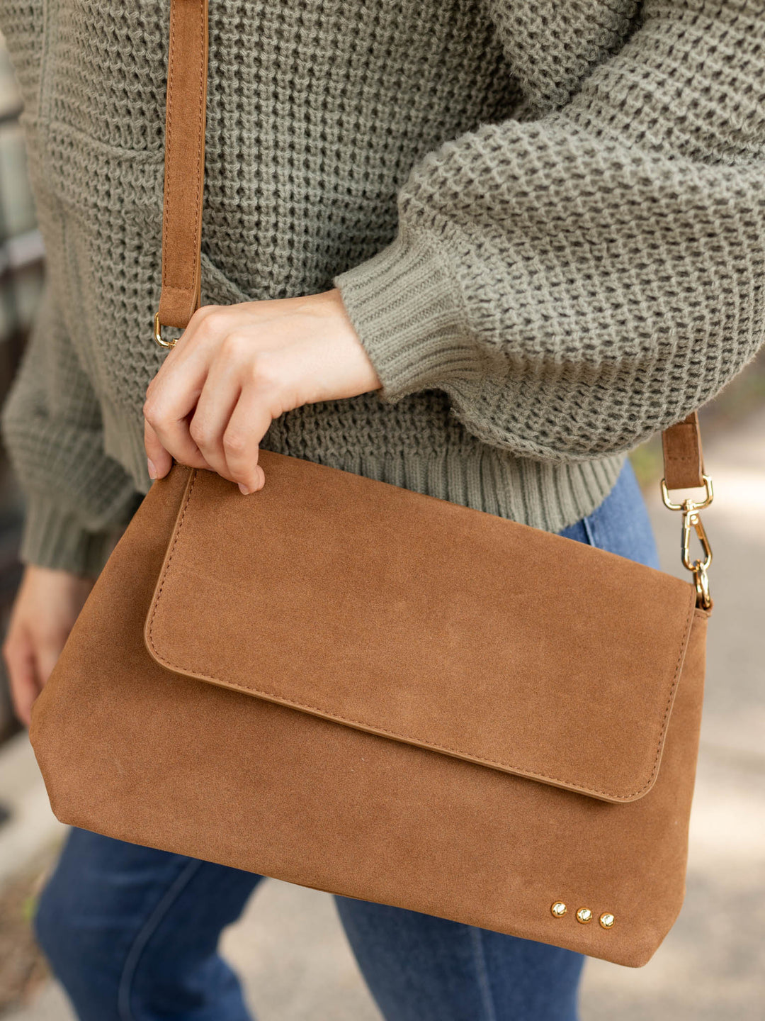 A person wearing a chunky knit green sweater and blue jeans carries a brown suede crossbody bag with a flap closure and gold-tone hardware.