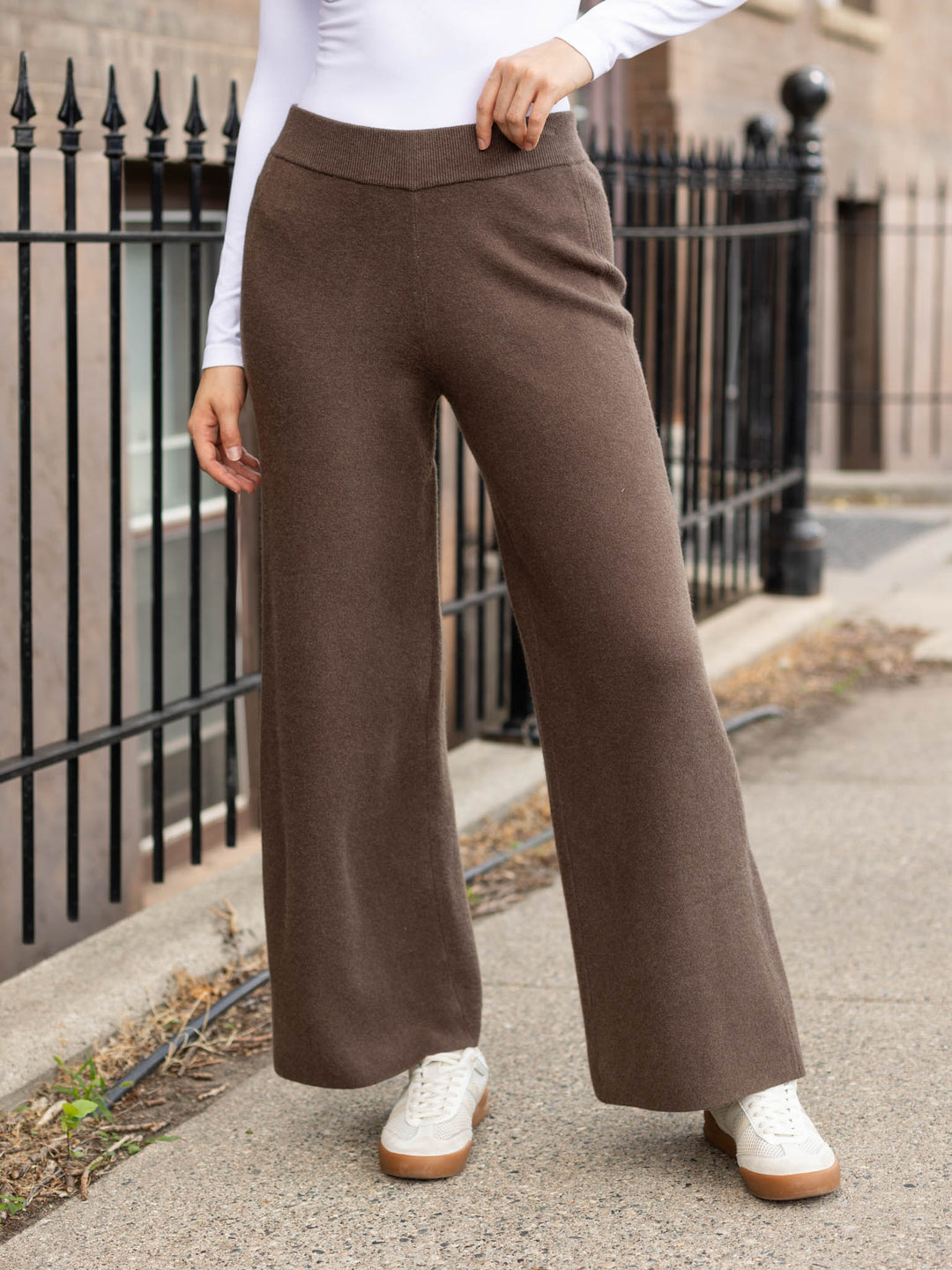 A person wearing high-waisted, wide-leg brown pants with a white long-sleeve top and white sneakers, standing outdoors on a sidewalk next to a black metal fence.
