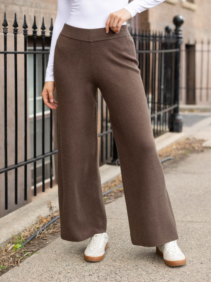 A person wearing high-waisted, wide-leg brown pants with a white long-sleeve top and white sneakers, standing outdoors on a sidewalk next to a black metal fence.