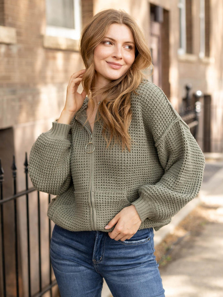 A woman wearing an olive green waffle-knit zip-up cardigan and blue jeans, standing outdoors on a city sidewalk in front of brick buildings.