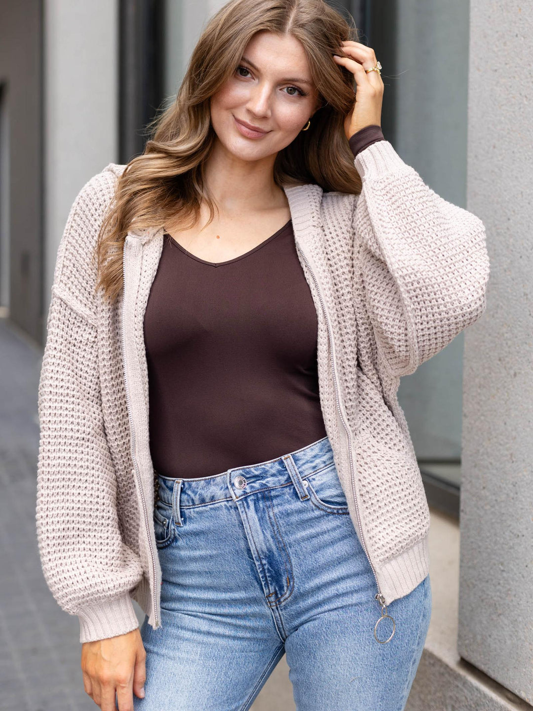 A woman wearing a light beige open-front knit cardigan over a dark brown camisole and blue jeans, standing outdoors against a gray building wall.