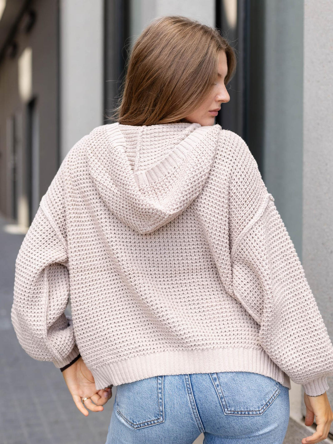 A woman viewed from behind wearing a chunky knit hooded sweater in a light blush pink color, paired with blue jeans, in an outdoor urban setting.