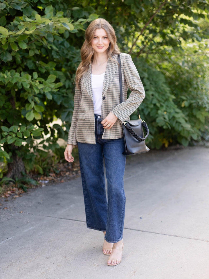 A woman stands outdoors on a paved path wearing a beige and brown houndstooth blazer, blue wide-leg jeans, a white top, and bare feet, while carrying a black shoulder bag.