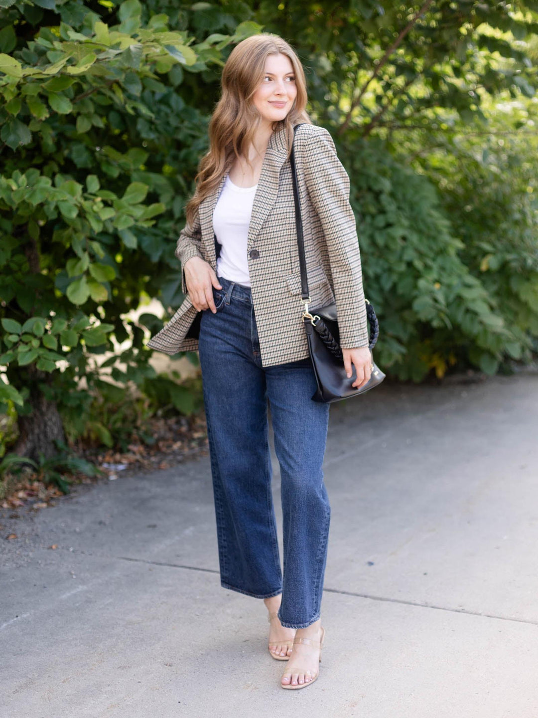 A woman stands outdoors on a paved path wearing a beige and brown houndstooth blazer, a white top, blue jeans, and clear high-heel sandals, while carrying a black shoulder bag.