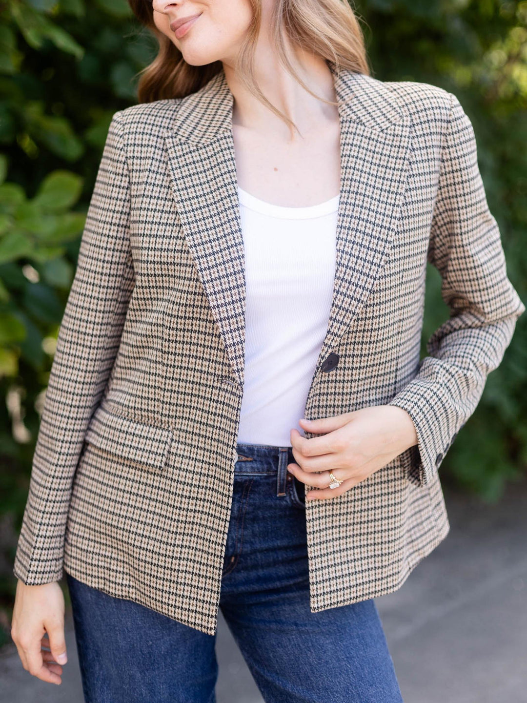 A woman wearing a beige and brown houndstooth blazer over a white tank top and blue jeans, standing outdoors with green foliage in the background.