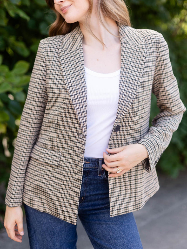 A woman wearing a beige and brown houndstooth blazer over a white tank top and blue jeans, standing outdoors with green foliage in the background.