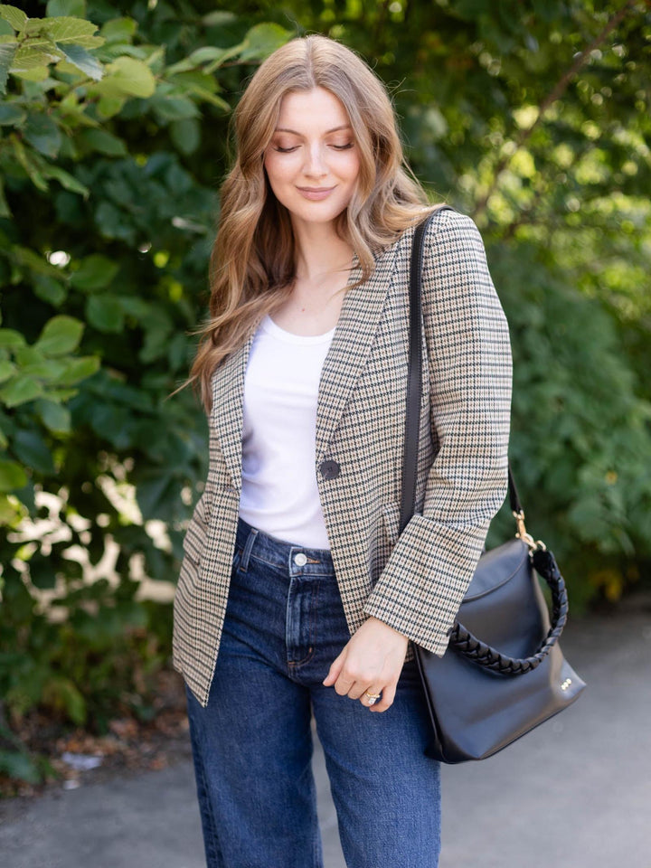 A woman wearing a beige and black checkered blazer over a white top and blue jeans, carrying a black shoulder bag with a braided strap, standing outdoors in front of green foliage.