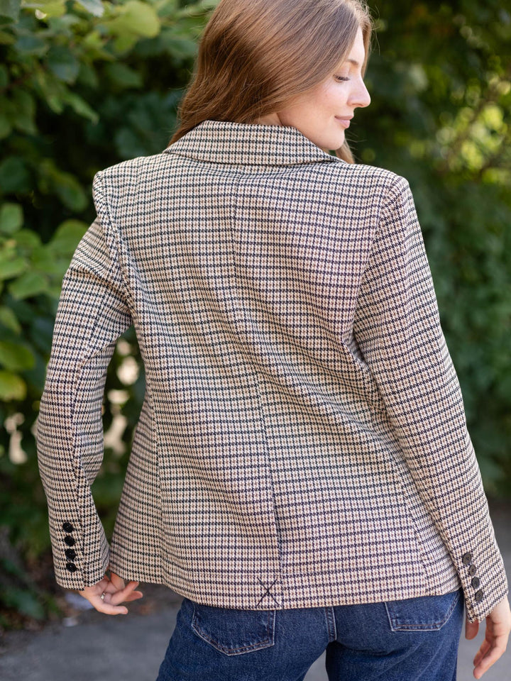 A woman wearing a beige and brown houndstooth blazer, shown from the back in an outdoor setting with green foliage in the background.