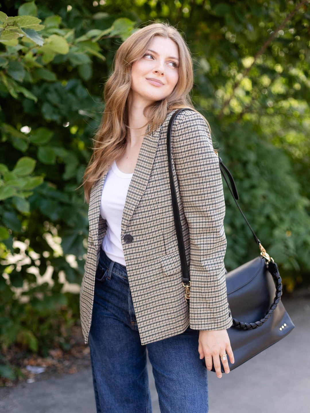 A woman wearing a beige and brown houndstooth blazer over a white top and blue jeans, carrying a black shoulder bag with a braided strap, set against a background of green foliage.