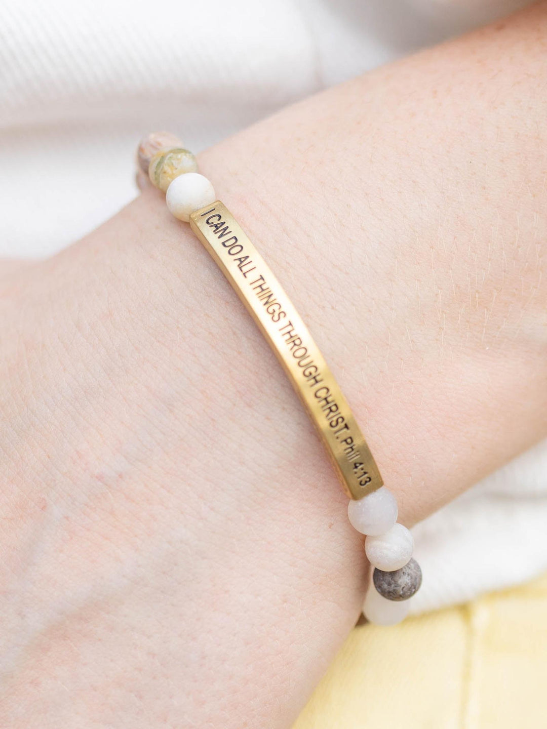 Stone bracelet with "I CAN DO ALL THINGS THROUGH CHRIST PHIL 4:13" inscribed on a gold bar, worn on a wrist.