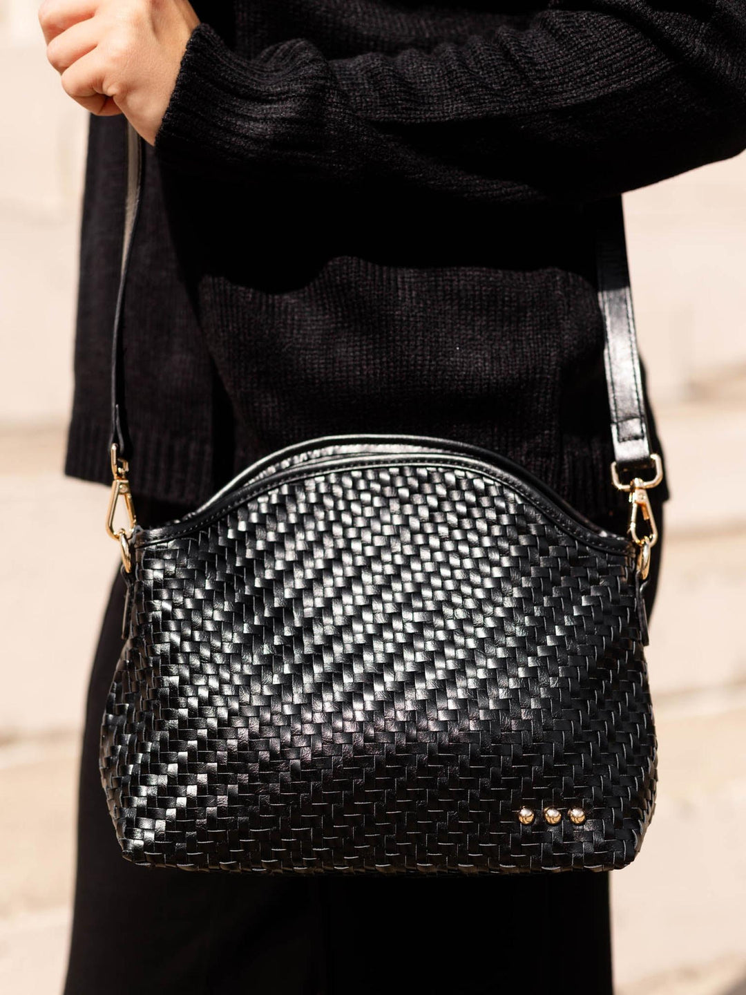 A black woven leather shoulder bag with gold-tone hardware, worn crossbody by a person in a black sweater against a light stone wall.