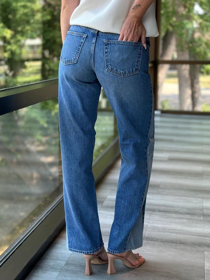 A person wearing blue wide-leg denim jeans, shown from the back while standing on a balcony with a glass railing and outdoor greenery in the background.