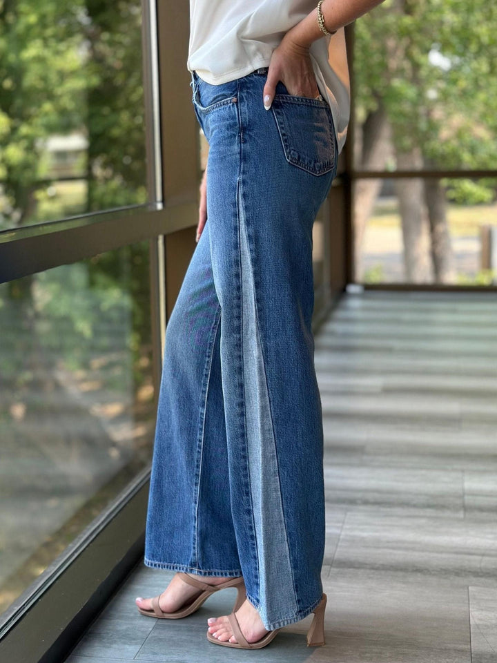 A person wearing wide-leg blue denim jeans with a front slit, paired with a white top and beige heeled sandals, standing on a balcony with a view of green trees.