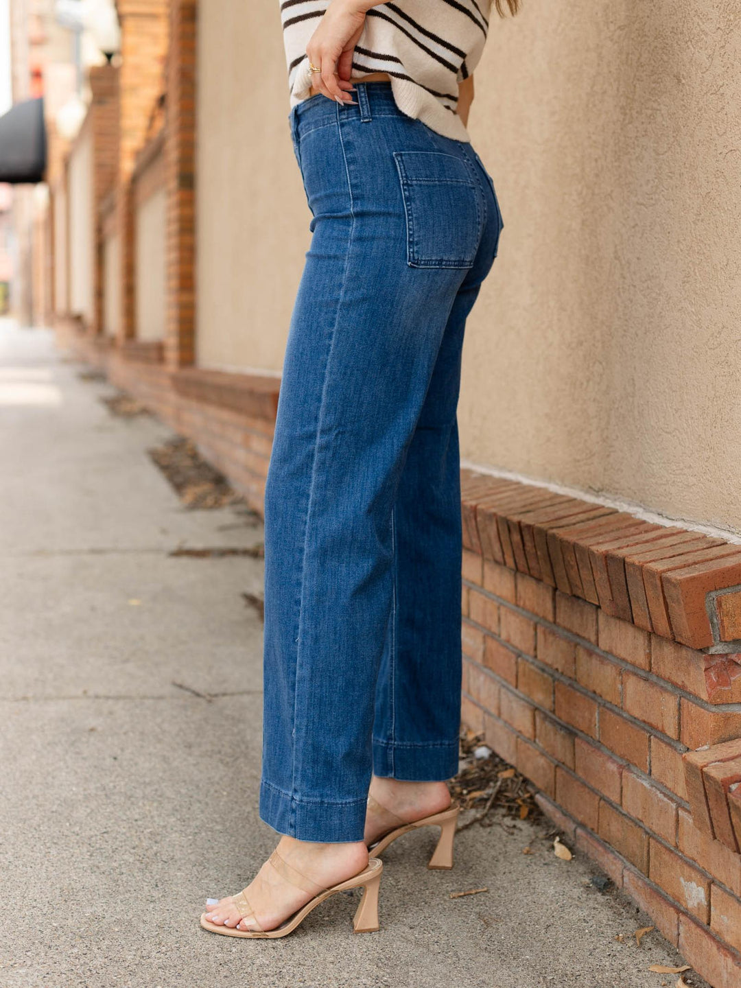 A person wearing blue wide-leg cropped jeans, a cream and black striped top, and beige heeled sandals, standing on a sidewalk next to a beige wall and brick base.