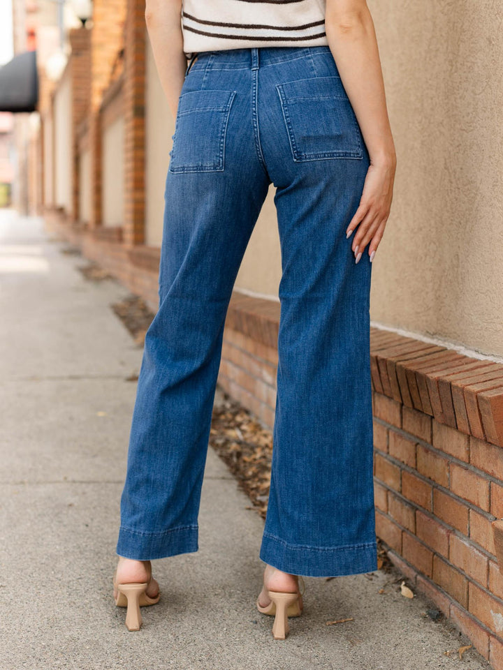 A person shown from the back wearing blue wide-leg jeans, a striped top, and beige heeled sandals, standing on a sidewalk beside a brick wall.