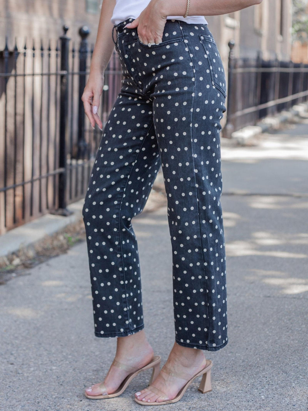 A person wearing black wide-leg cropped pants with a white polka dot pattern, paired with a white top and clear strap block-heel sandals, standing on an outdoor paved street.