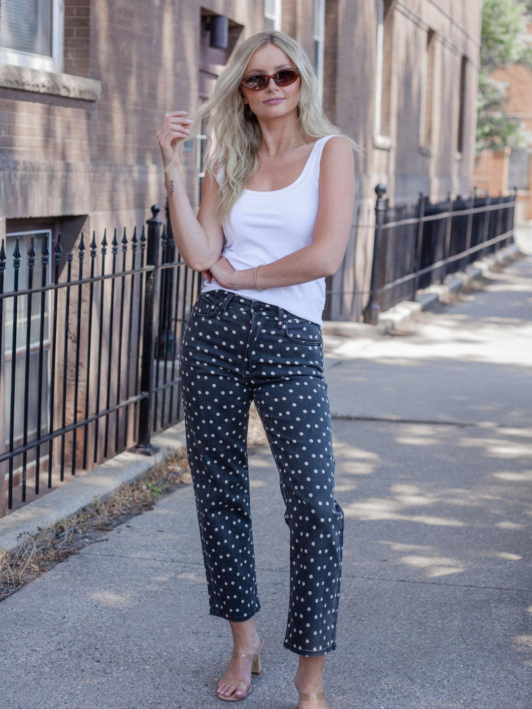 A woman stands on a city sidewalk wearing dark navy pants with a white polka dot pattern, a white tank top, and sunglasses.