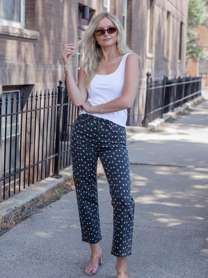 A woman stands on a city sidewalk wearing dark navy pants with a white polka dot pattern, a white tank top, and sunglasses.