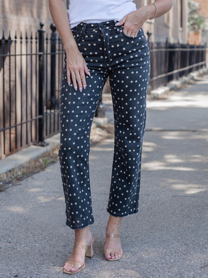 A person wearing dark navy cropped pants with a white polka dot pattern, styled with a white top and clear high-heeled sandals on an outdoor sidewalk.