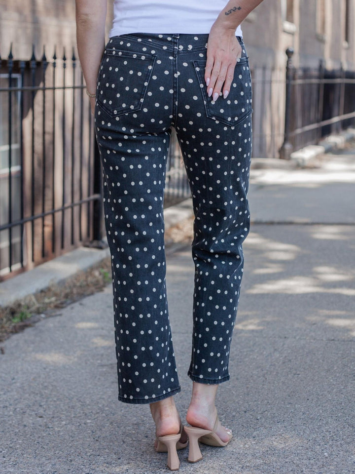 A person shown from the back wearing dark navy pants with a white polka dot pattern, paired with a white top and beige heeled sandals, standing on an outdoor paved walkway.
